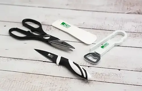 Ein Messer in Schwarz-Weiß, eine schwarze Schere, ein weißer Spatel mit dem P&T-Logo in Grün und ein weißer Flaschenöffner mit dem P&T-Logo liegen auf einem Tisch.