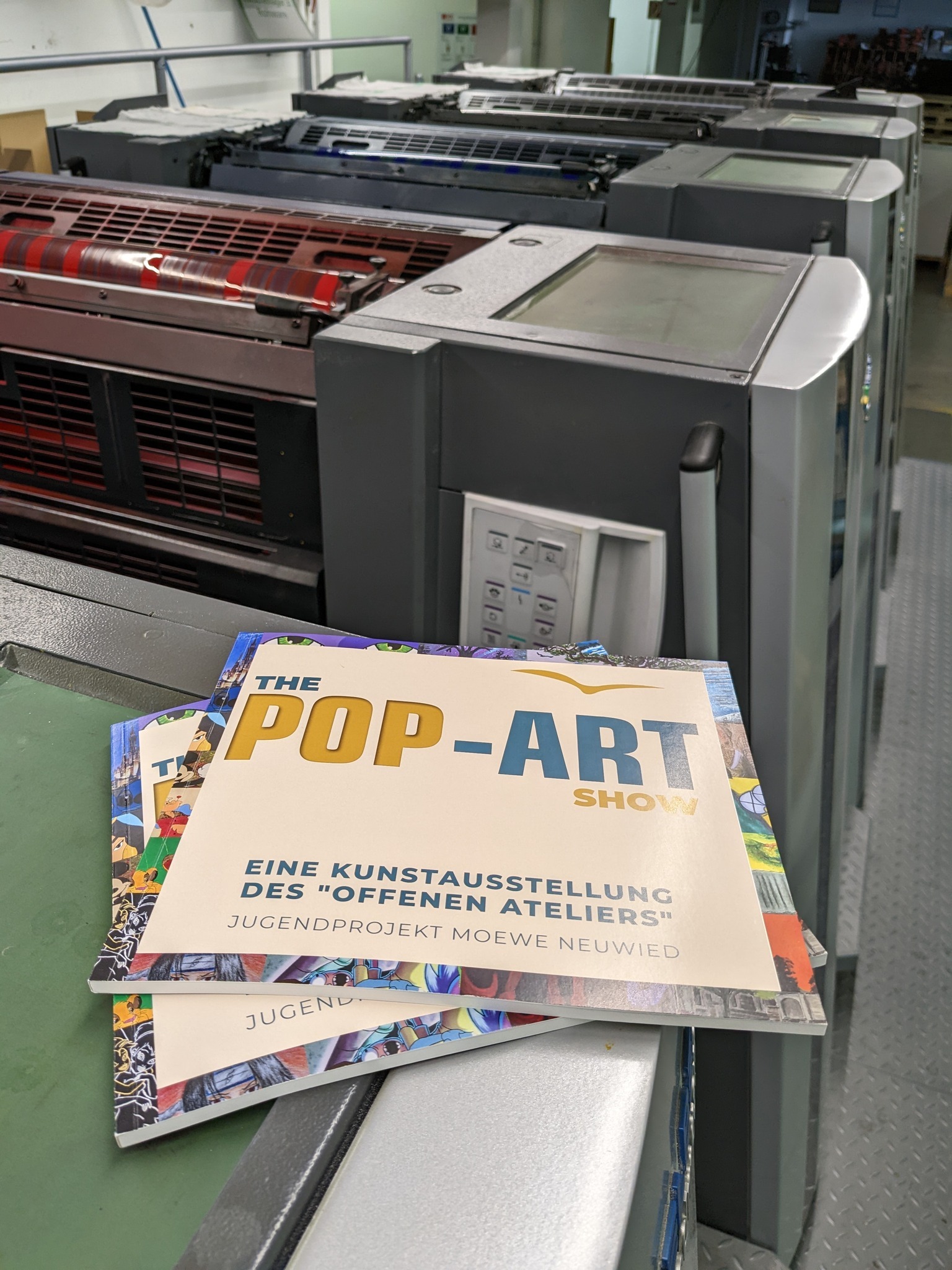 Eine Zeitung, die zur Hälfte zu sehen ist, mit blauer und gelber Schrift: ‚The Pop Art Show‘. Im Hintergrund sieht man einen Teil einer Druckermaschine