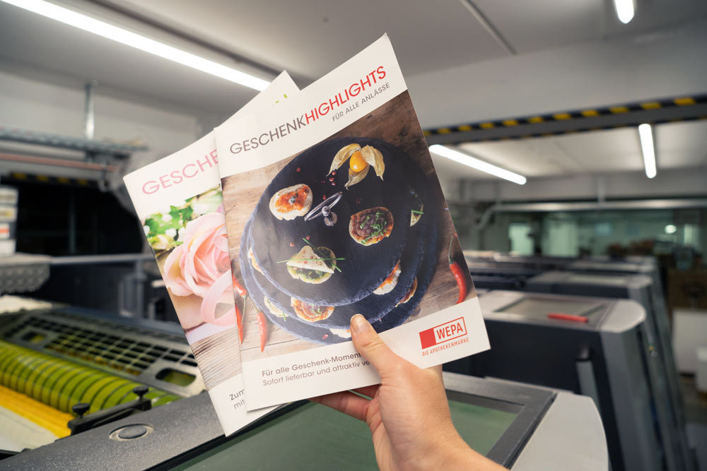 Eine Hand, die zwei Wepa-Kataloge hochhält, mit dem Titel 'Geschenke-Highlights'. Im Hintergrund sind unscharf Druckmaschinen zu erkennen.